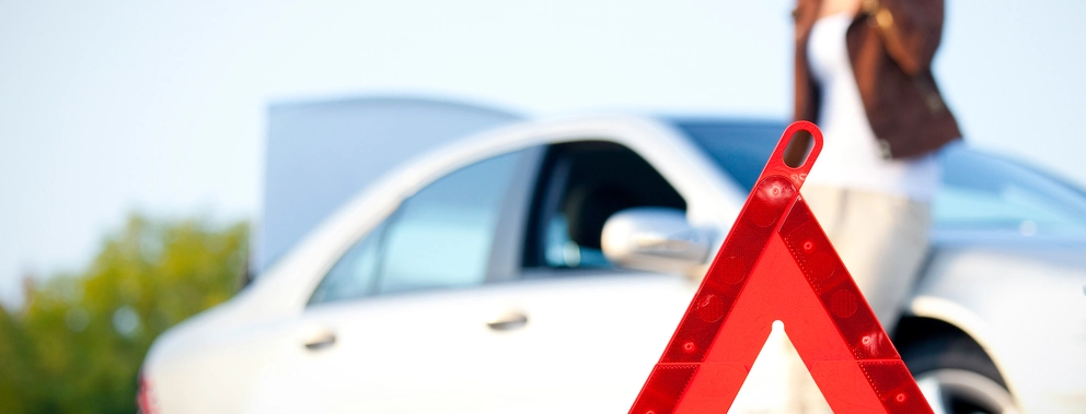 Im Vordergrund steht ein rotes Warndreieck auf der Strasse. Im Hintergrund ist ein weisses Auto mit geöffneter Motorhaube zu sehen, daneben eine unscharfe Frau, die telefoniert. Die Szene deutet auf eine Panne hin.