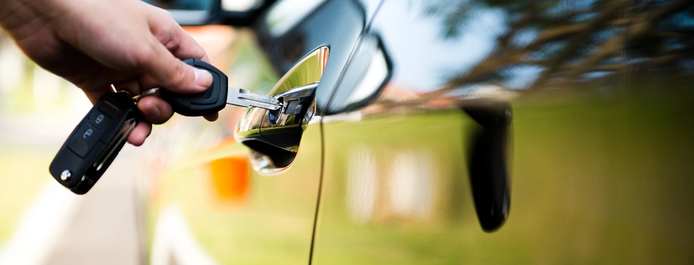 Nahaufnahme einer Hand, die einen Autoschlüssel in das Schloss eines glänzenden, schwarzen Autos steckt. Die Autotür und der Hintergrund spiegeln die Umgebung mit unscharfem Gras und Bäumen wieder.